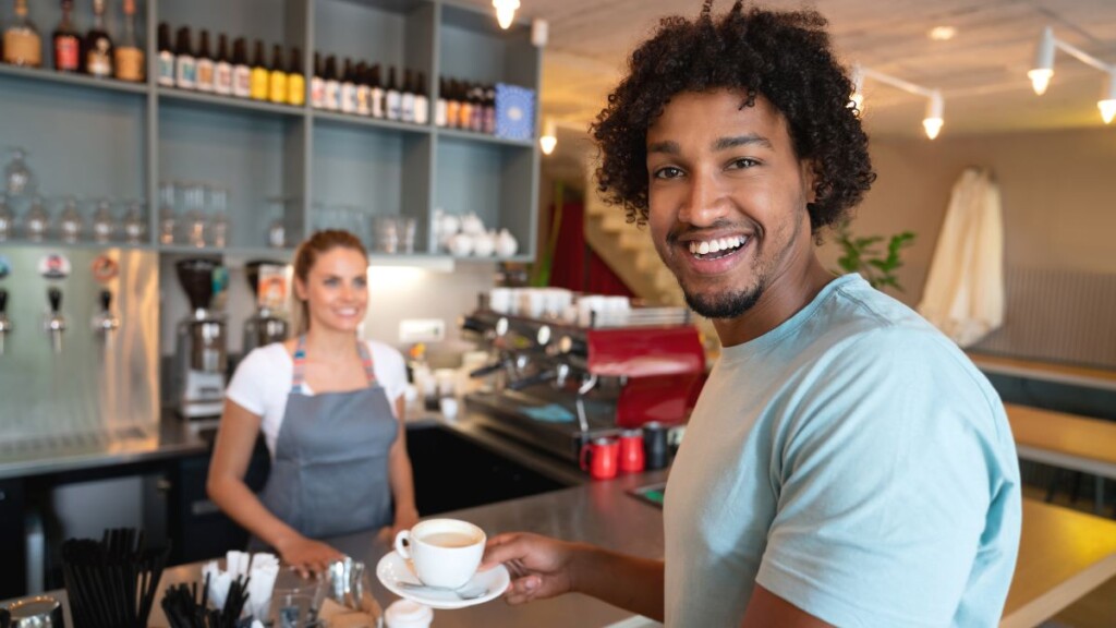 Photo of a coffeeshop customer smiling at the camera as a barista hands him a cup of coffee